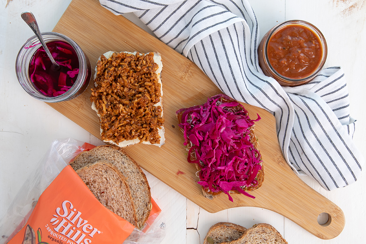 Sweet & Spicy BBQ Shredded Tofu on a slice of Full Seed Ahead sprouted bread with hummus. The other half of the sandwich with Quick-Pickled Red Onions sits beside it on a cutting board.