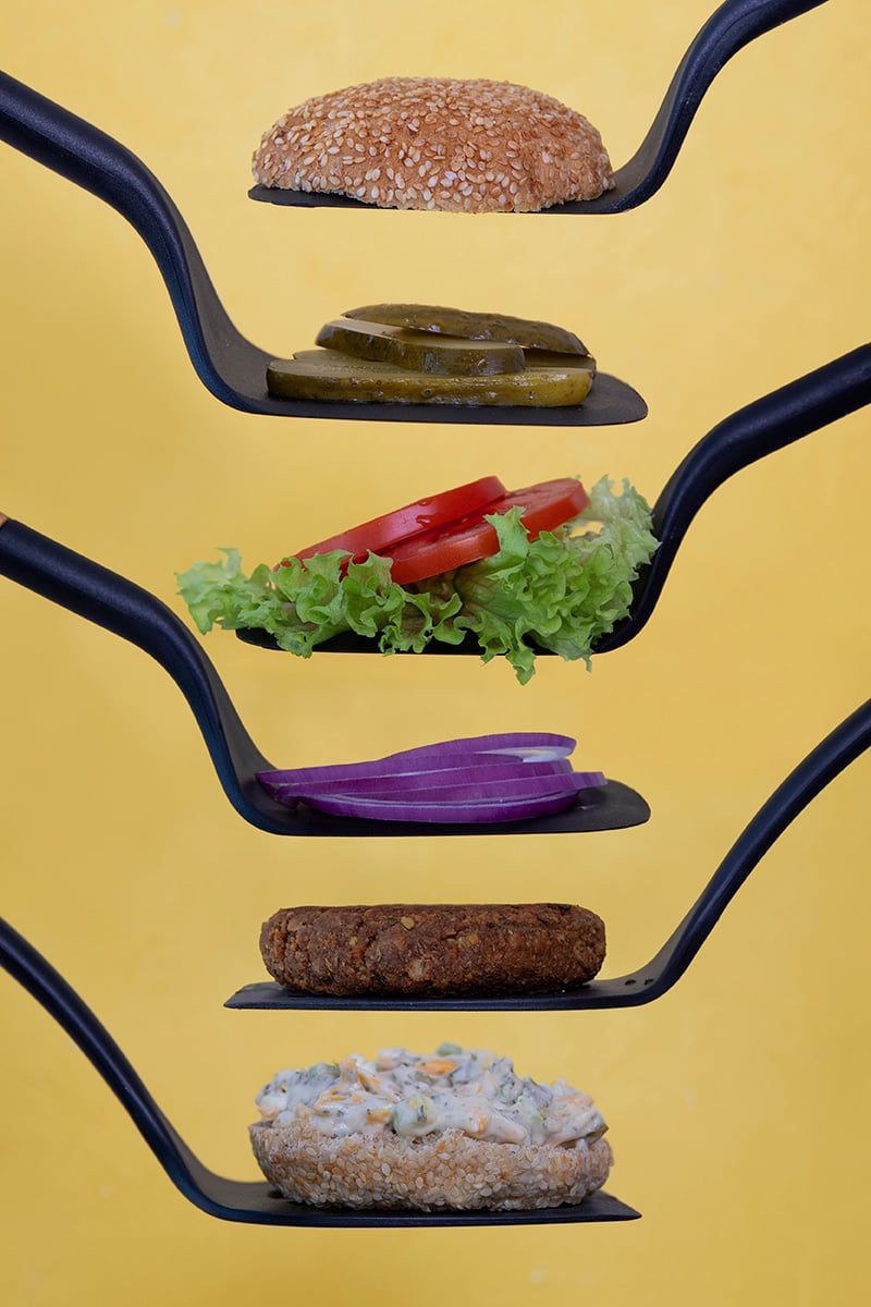 Six spatulas showing each layer of a Sprouted Green Lentil Burger: [from the top] the top of a Silver Hills Bakery sprouted whole grain Sesame Burger Bun; sliced pickles; lettuce and tomato; sliced red onion; Sprouted Green Lentil veggie burger patty; bottom bun with with Vegan Pickle Dip Ranch Sauce